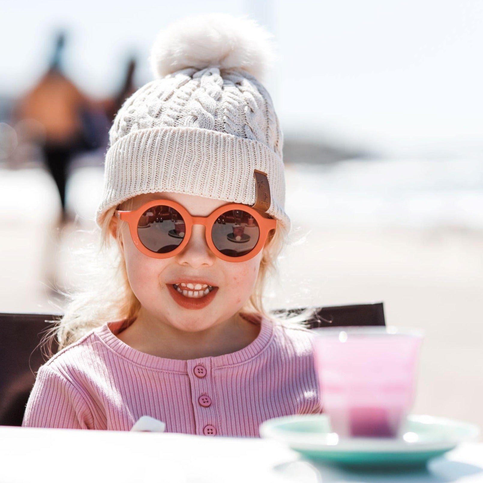 Girl wearing cream pom pom beanie and kids orange terracotta sunglasses with UV protection. Cubs & Co. Australia