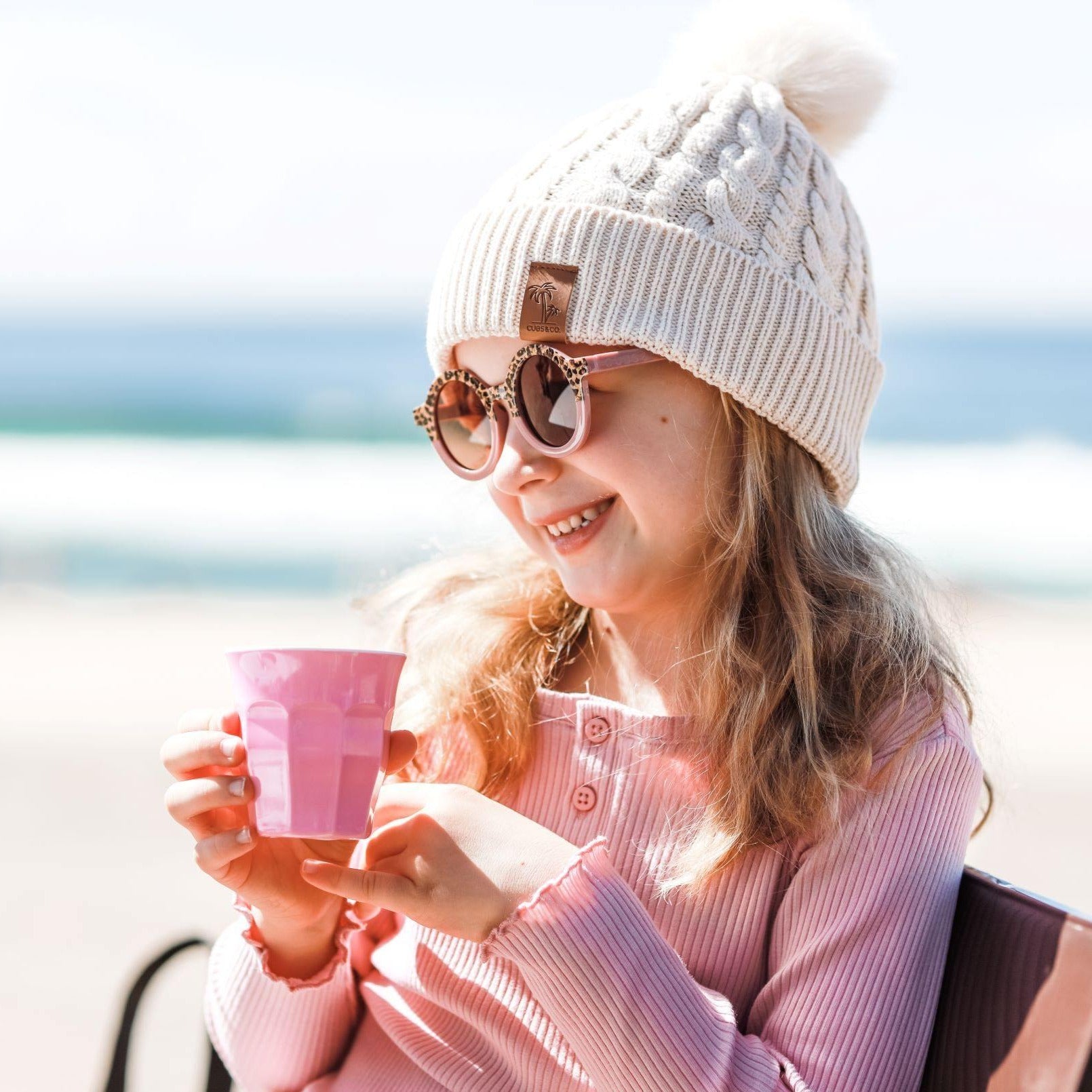 Girl wearing cream pom po0m beanie and kids pink leopard sunglasses with UV protection. Cubs & Co. Australia